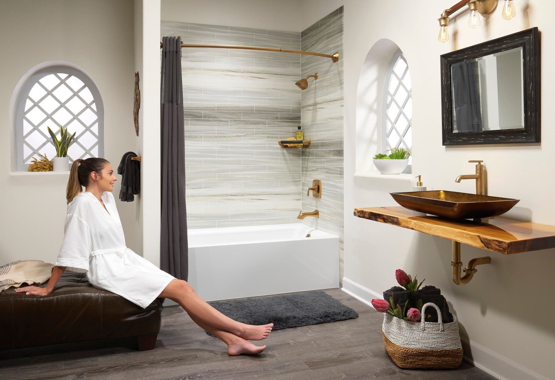 Bright, spa-inspired bathroom featuring a white bathtub with tiled surround, brushed gold fixtures, floating wood vanity with vessel sink, decorative arched windows, and a woman in a robe relaxing in the modern, inviting space.