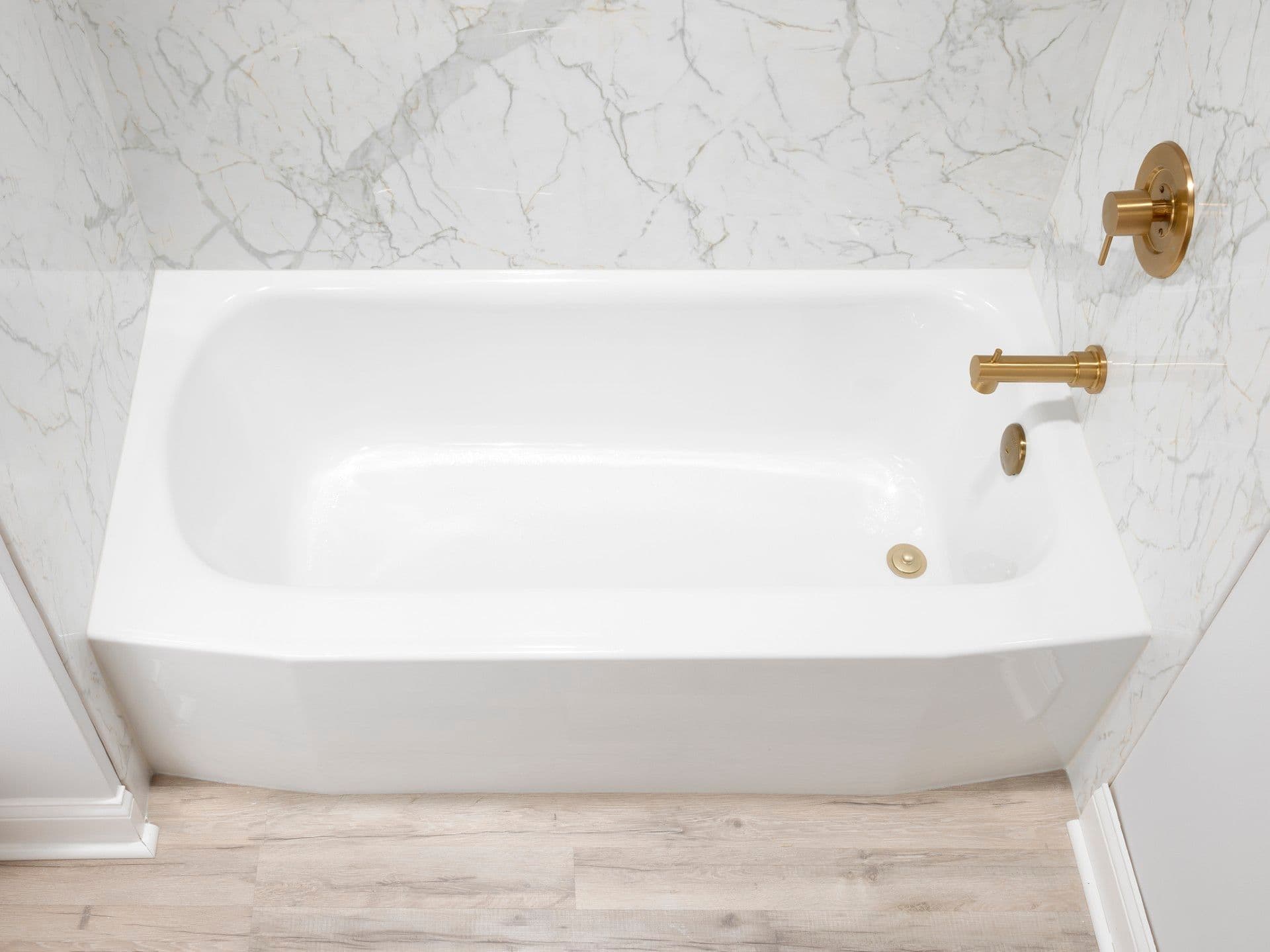 Overhead view of a sleek white bathtub with a marble-look wall surround, brushed gold fixtures, and light wood-look flooring for a clean, modern bathroom design.