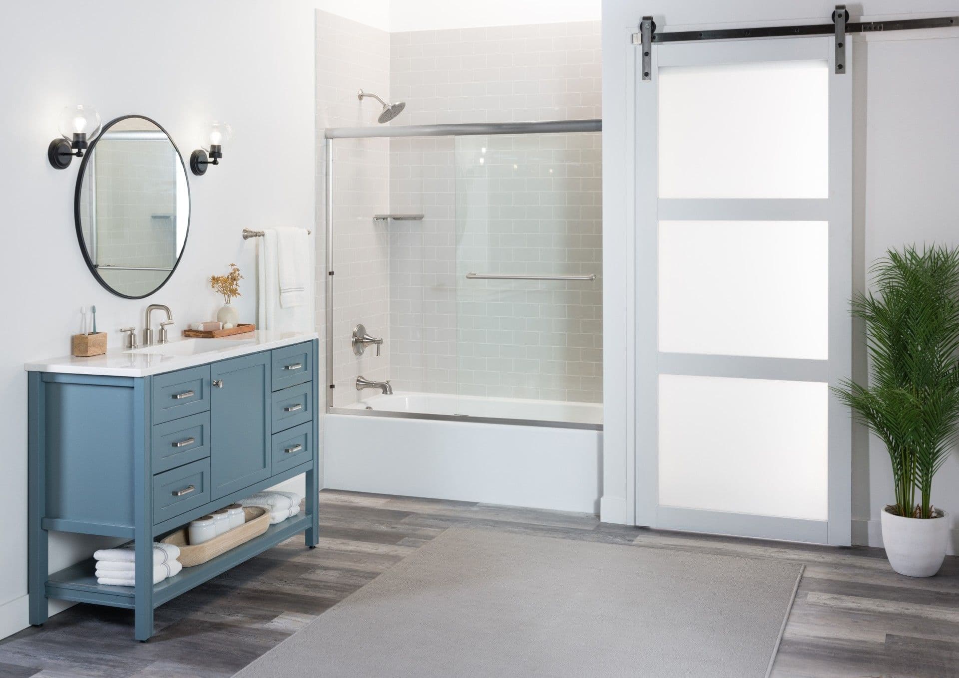 Bright, modern bathroom with a blue vanity, round black-framed mirror, white subway tile tub surround, sliding glass tub enclosure, gray wood-look flooring, and a contemporary barn-style sliding door.