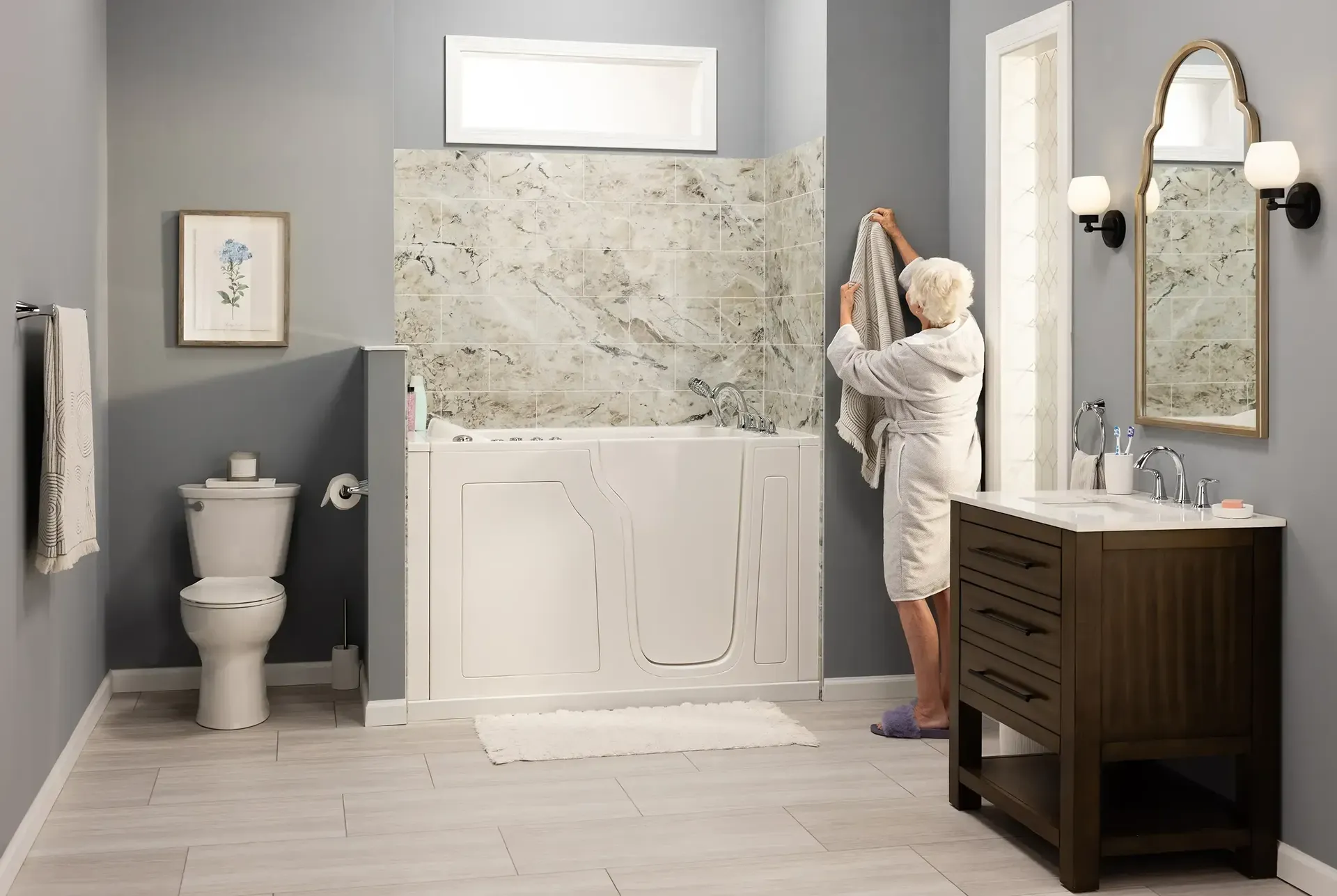 Bright, accessible bathroom featuring a walk-in bathtub with a low-entry door, marble-look tile surround, modern wood vanity with quartz countertop, and an older adult in a robe hanging a towel, highlighting comfort, safety, and independent living.