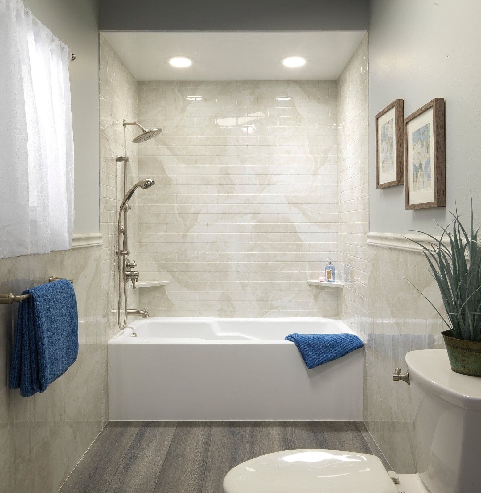 Modern bathroom with a low-threshold walk-in bathtub, marble-look tiled shower surround, dual showerheads with handheld sprayer, built-in corner shelves, recessed lighting, and wood-look flooring designed for comfort, safety, and accessibility.